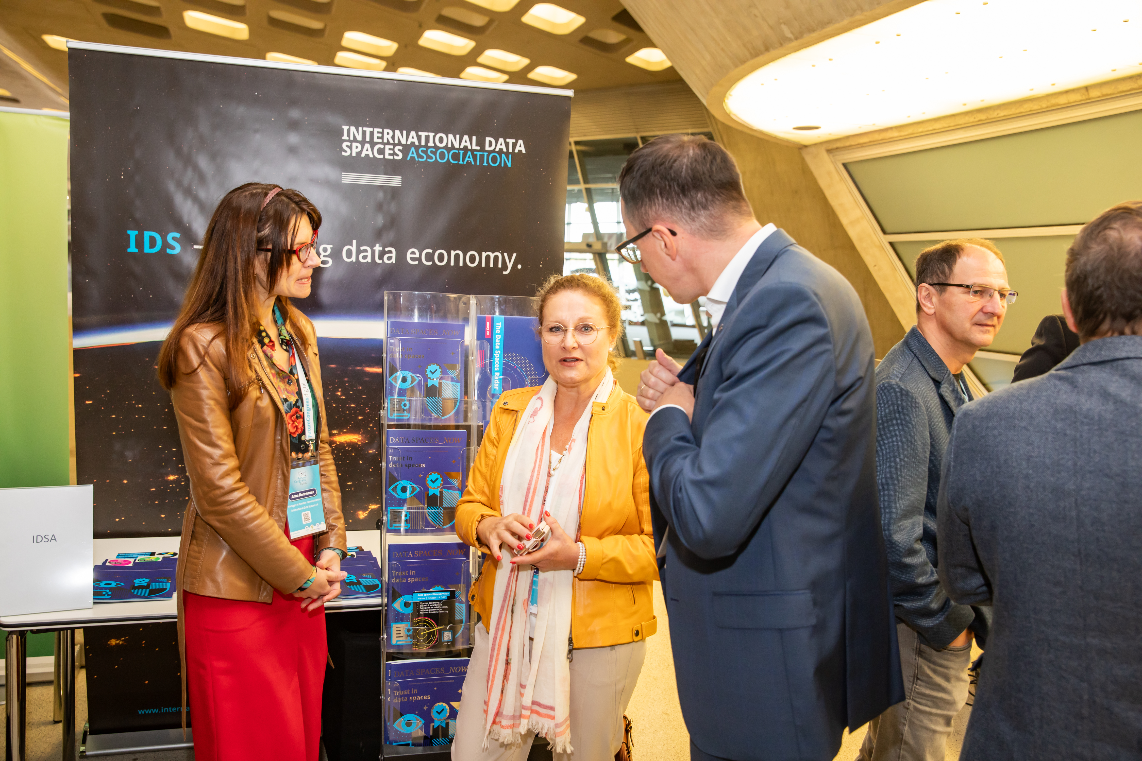 Menschen unterhalten sich vor dem Stand von der International Data Spaces Association. Im Hintergrund befinden sich ein großes Roll-Up und Magazine.