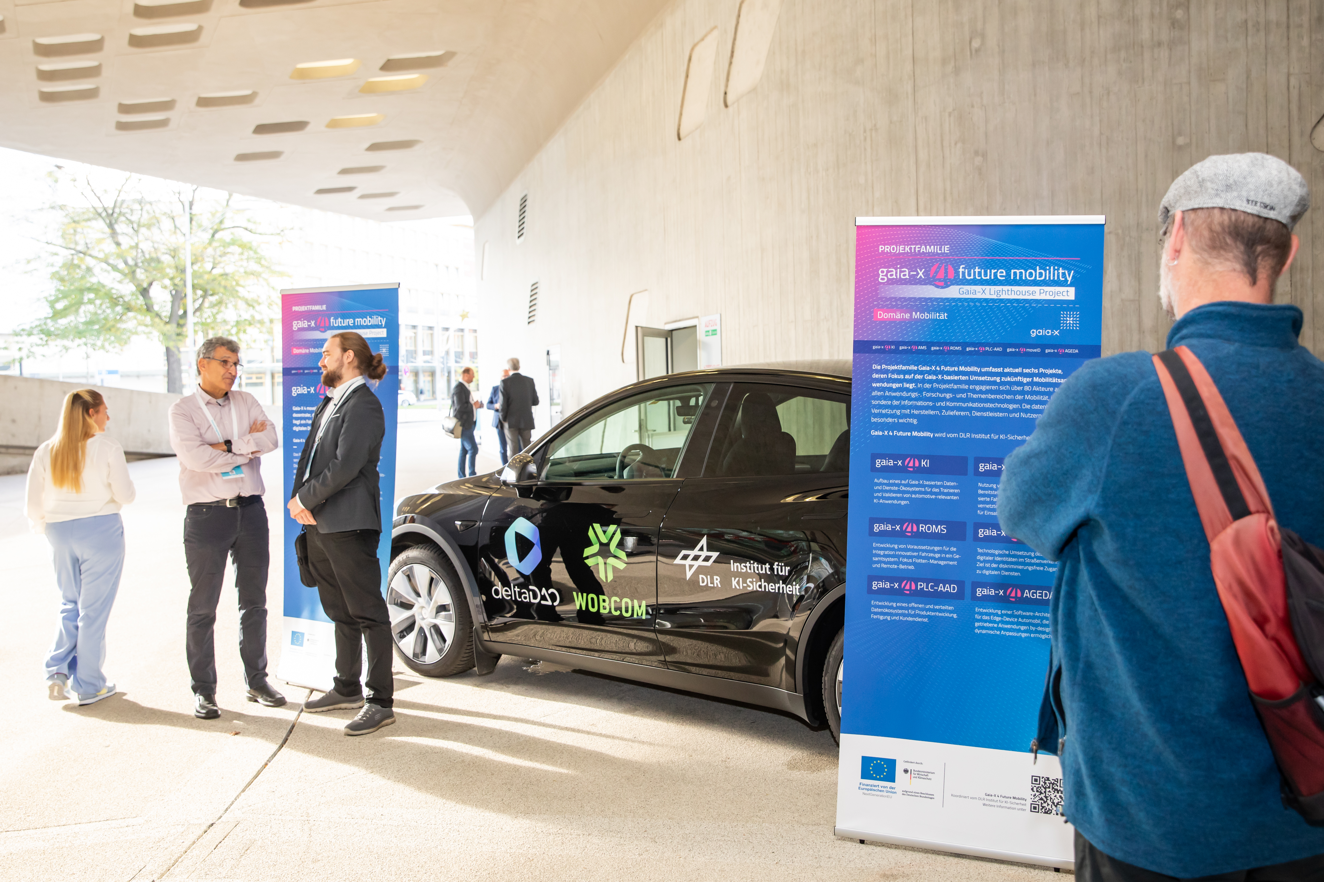 Menschen vor dem Stand von gaia-x. In der Mitte steht ein Auto mit den Logos von deltaDAO, WOBCOM und dem Institut für DLR Institut für KI-Sicherheit. Links und rechts stehen zwei Roll-Ups mit weiterführenden Informationen.